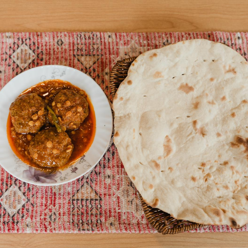 Three lamb meatballs in orange gravy in a flowered bowl beside a big round naan in a basket, on a carpet runner