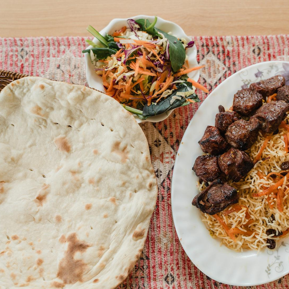 Grilled lamb pieces on Qabuli rice with carrots and raisins, flatbread and a fresh salad on a carpet runner