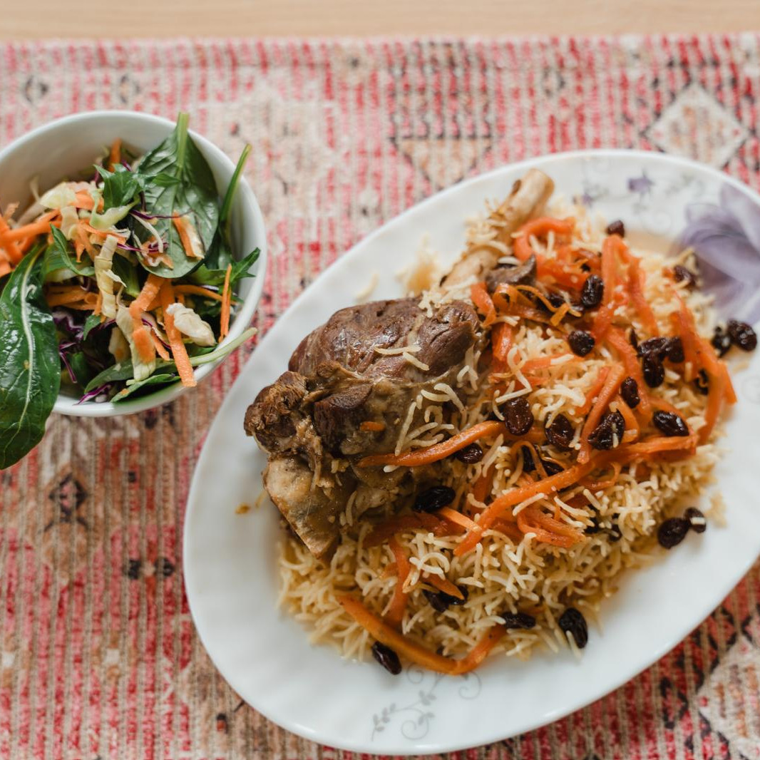 Lamb shank on Qabuli rice with raisins and carrots, salad beside it, on a red Afghan carpet runner